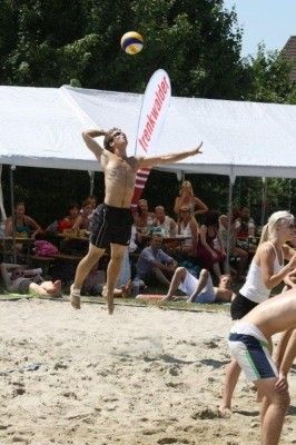 Ein Mann springt, um einen Volleyball auf einem Sandstrand zu schlagen, während Zuschauer unter einem weißen Zelt zusehen.
