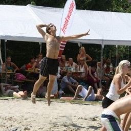 Ein Mann springt, um einen Volleyball auf einem Sandstrand zu schlagen, während Zuschauer unter einem weißen Zelt zusehen.