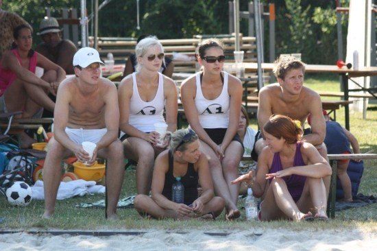 Eine Gruppe von Menschen sitzt auf dem Gras, einige tragen weiße Tanktops mit der Nummer 1, während andere oberkörperfrei sind. Sie beobachten wahrscheinlich ein Spiel.