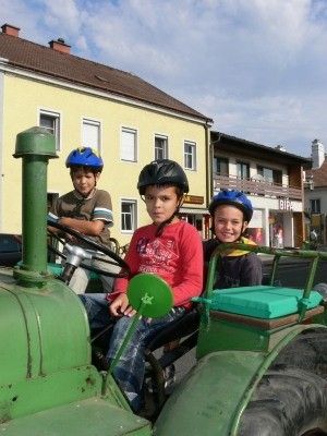 Drei Kinder fahren auf einem grünen Traktor vor einem gelben Gebäude mit einem Schild, das 'BIP' steht. Die Kinder tragen Helme und scheinen für ein Foto zu posieren.