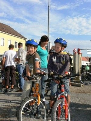 Zwei junge Jungen mit Helmen stehen neben ihren Fahrrädern an einem sonnigen Tag, mit mehreren Menschen und Fahrrädern im Hintergrund.