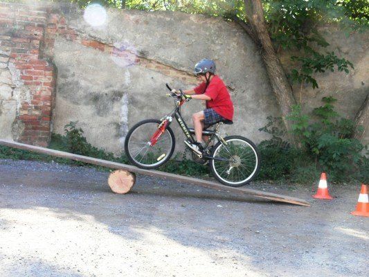 Ein Kind fährt mit einem Fahrrad eine Holzrampe in einem Park hinauf, mit orangefarbenen Verkehrskegel in der Nähe.