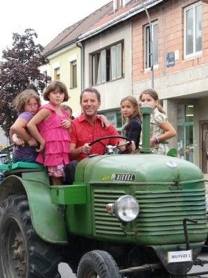 Ein Mann fährt einen grünen Traktor mit vier Mädchen auf dem Schoß vor einem Gebäude.