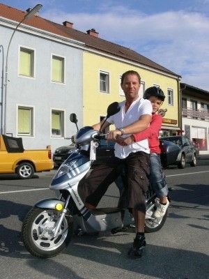 Ein Mann fährt mit einem Kind auf dem Schoß einen Roller in einer Stadtstraße mit Gebäuden und geparkten Autos im Hintergrund.