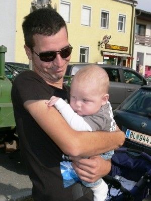Ein Mann mit Sonnenbrille hält ein schlafendes Baby in den Armen auf einem Parkplatz. Hinter ihnen sind Gebäude und geparkte Autos zu sehen.