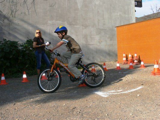 Ein junger Junge mit Helm fährt ein Fahrrad durch einen Parcours, der von orangefarbenen Verkehrskegeln markiert ist, auf einem Kiesweg. Eine Frau steht in der Nähe und hält eine Kamera.