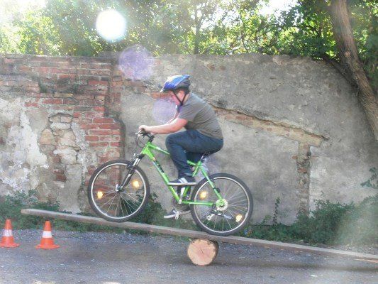 Ein Mann fährt ein grünes Fahrrad auf einem Holzbrett, das auf einem Baumstamm balanciert, trägt einen Helm und Turnschuhe. Er steht vor einer Ziegelmauer mit einem Baumstamm auf der rechten Seite.