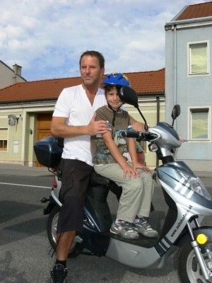 Ein Mann und ein junger Junge fahren einen Roller auf einer Straße vor einem Gebäude. Der Junge trägt einen Helm.