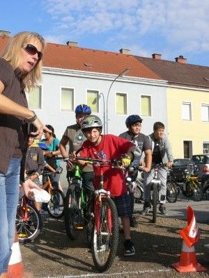 Eine Frau mit Sonnenbrille und einem braunen Shirt steht neben einer Gruppe von Kindern mit Helmen, die Fahrräder in einem Parkplatz fahren. Im Hintergrund sind Gebäude und Autos zu sehen.