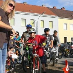 Eine Frau mit Sonnenbrille und einem braunen Shirt steht neben einer Gruppe von Kindern mit Helmen, die Fahrräder in einem Parkplatz fahren. Im Hintergrund sind Gebäude und Autos zu sehen.