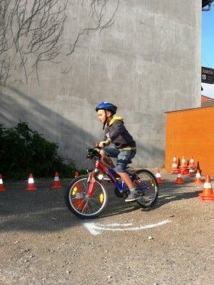 Ein junger Junge fährt auf einem Kiesweg mit einem roten Fahrrad und einem blauen Helm. Er navigiert durch eine Reihe von orangefarbenen Verkehrskegeln.