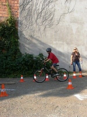 Ein junger Junge fährt mit einem Fahrrad durch eine Reihe von orangefarbenen Hütchen auf einem Kiesweg. Eine Frau steht in der Nähe und beobachtet.
