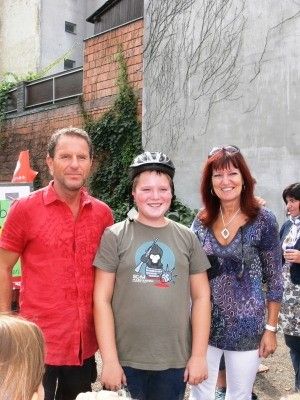 Drei Personen stehen zusammen draußen in der Nähe eines Gebäudes, ein Mann in einem roten Shirt, ein Junge in einem grauen Shirt mit Helm und eine Frau in einem blauen Shirt.