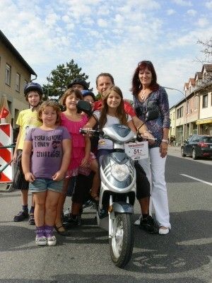Eine Familie mit sieben Personen, darunter Kinder, steht neben einem Roller auf einer Straße, mit Gebäuden und Bäumen im Hintergrund.