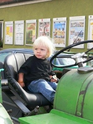 Ein junges Kind mit blonden Haaren sitzt auf dem Fahrersitz eines grünen Traktors, mit einem Gebäude und mehreren Postern im Hintergrund.