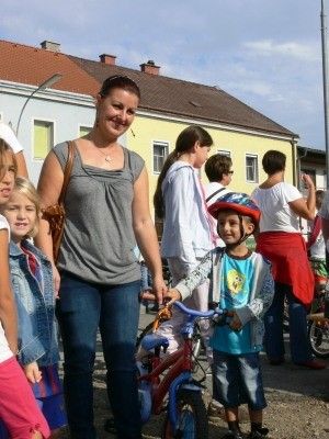 Eine lächelnde Frau steht mit einem Kind, das ein Fahrrad hält, an einem Außenbereich. Einige Leute stehen hinter ihnen, und einige gehen auf der Straße. Ein Gebäude befindet sich hinter ihnen.