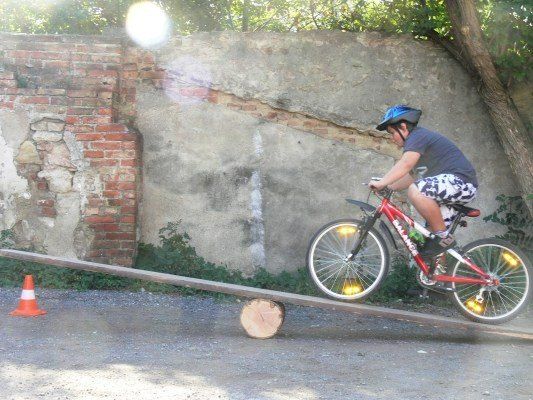 Ein junger Junge fährt auf einem rot lackierten Fahrrad auf einer Wippe und balanciert auf einem Baumstamm, während er einen Helm trägt. Die Szene spielt sich vor einer Backsteinmauer mit Grünpflanzen ab.