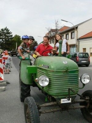 Ein Mann fährt einen grünen Traktor mit drei Kindern darauf. Sie tragen Helme und scheinen die Fahrt zu genießen. Im Hintergrund befinden sich Gebäude, ein geparktes Auto und gehende Menschen.