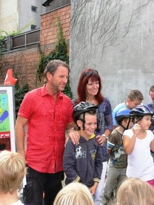 Ein Mann in einem roten Shirt und eine Frau in einem blauen Shirt stehen mit Kindern vor einem Gebäude. Die Kinder tragen Helme und lächeln.
