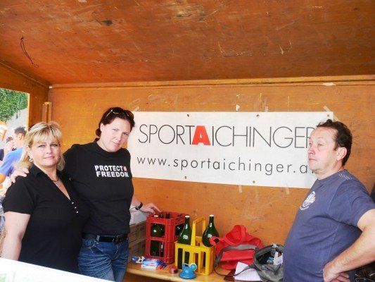 Drei Personen stehen vor einem Stand mit einem Banner, das 'SPORT AICHINGER' besagt. Die Frau links trägt ein schwarzes Shirt mit 'PROTECT FREEDOM', die mittlere Person trägt ein schwarzes Shirt mit Sonnenbrille auf dem Kopf und der Mann rechts trägt ein blaues Shirt.