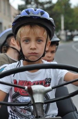 Ein junger Junge mit einem weißen T-Shirt mit Grafikdesign und einem blauen Helm sitzt auf einem Tretfahrzeug und sieht ernst aus. Hinter ihm sitzen zwei weitere Kinder, die ebenfalls Helme tragen.