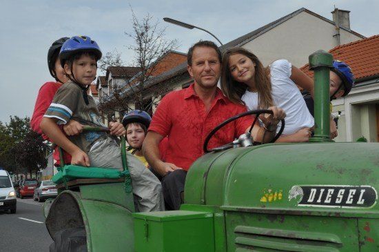 Ein Mann fährt einen grünen Traktor mit vier Kindern darauf. Sie tragen alle Helme. Der Mann trägt ein rotes Hemd. Hinter ihnen sind Gebäude und Autos auf der Straße.