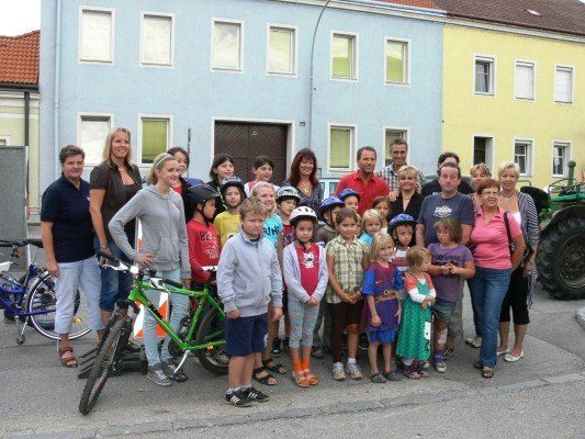 Eine Gruppe von Erwachsenen und Kindern, einige mit Helmen, stehen vor einer Reihe von Häusern. Fahrräder sind am Straßenrand geparkt.