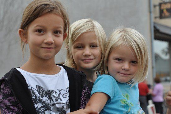 Drei junge Mädchen mit blonden Haaren stehen eng beieinander und lächeln für ein Foto. Das Mädchen links trägt ein weißes T-Shirt mit einem Schmetterlingsdesign, das mittlere ein schwarzes T-Shirt und das rechte ein blaues T-Shirt. Hinter ihnen ist ein Gebäude mit einem Schild zu sehen.