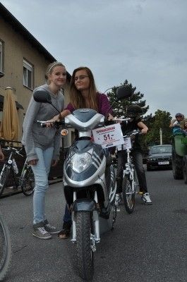 Zwei junge Frauen stehen neben einem Roller mit einem Nummernschild 51, in einer Außenumgebung mit Fahrrädern und einem Gebäude im Hintergrund.