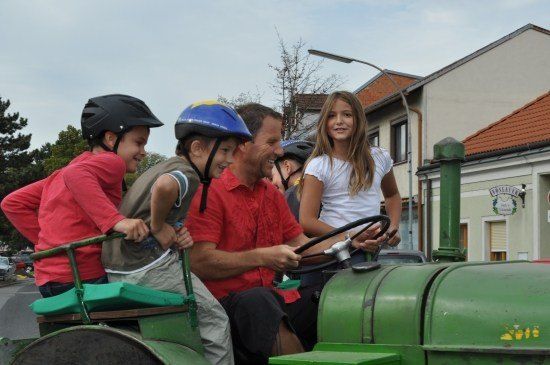 Ein Mann fährt einen grünen Traktor mit drei Kindern und einem Mädchen auf seinem Schoß. Sie tragen alle Helme.