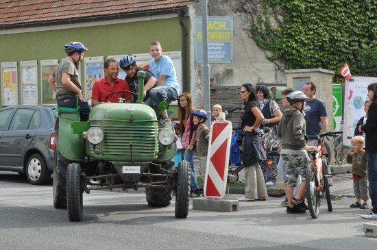 Eine Gruppe von Menschen fährt auf einem grünen Traktor auf einer Straße, mit mehreren Schaulustigen in der Nähe. Einige tragen Helme, und eine Frau hält ein Baby. Ein Auto und ein Fahrrad sind in der Nähe geparkt.