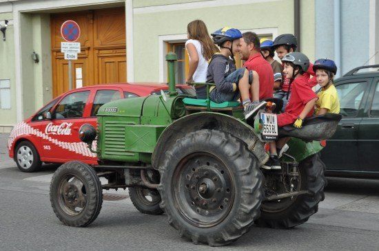 Eine Gruppe von Kindern und Erwachsenen fährt auf einem grünen Traktor auf einer Straße, mit einem roten Auto und einem Gebäude im Hintergrund.