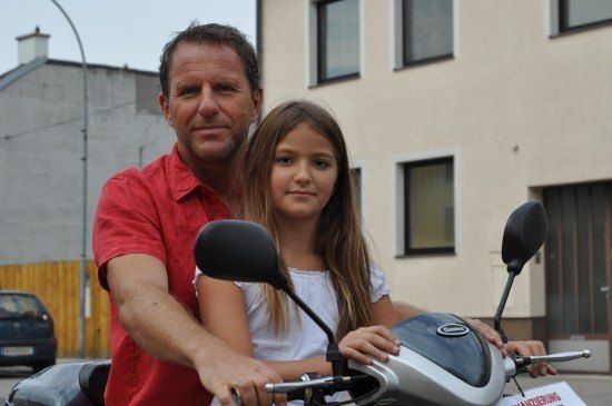 Ein Mann und ein junges Mädchen sitzen lächelnd auf einem Roller, mit einem Gebäude und einem geparkten Auto im Hintergrund.