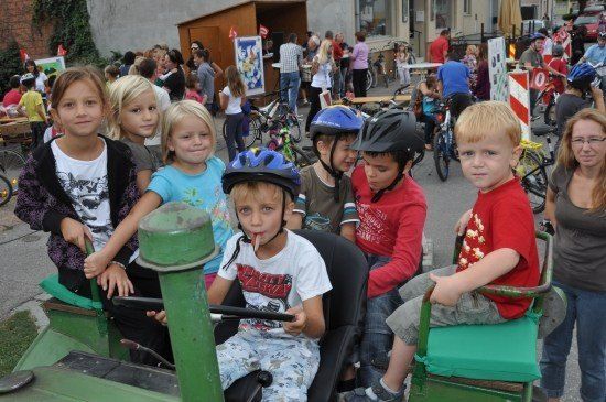 Eine Gruppe von Kindern sitzt auf einem grünen Traktor, trägt Helme und posiert für ein Foto bei einer Veranstaltung im Freien. Dahinter stehen und sitzen Menschen auf Fahrrädern.