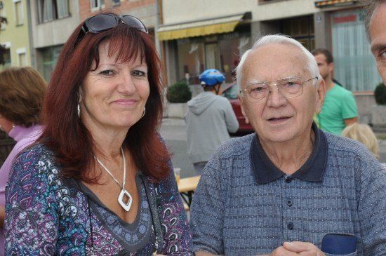Ein älterer Mann und eine Frau stehen gemeinsam auf einem Stadtplatz und lächeln für die Kamera. Hinter ihnen sind eine Person mit blauem Helm und eine weitere in einem grünen Shirt zu sehen.