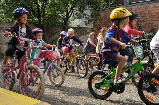 Eine Gruppe junger Kinder, alle mit Helmen, fahren auf einem Kiesweg mit ihren Fahrrädern. Sie stehen in einer Reihe, einige tragen Turnschuhe und andere Sandalen. Bäume und eine Ziegelmauer sind im Hintergrund zu sehen.
