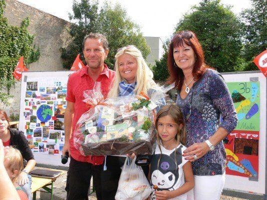Eine Gruppe von Menschen, darunter ein Mann, eine Frau und ein Kind, stehen zusammen und halten einen Geschenkkorb und Plastiktüten, vor einem Hintergrund von einer Wand mit Bildern und Pflanzen.