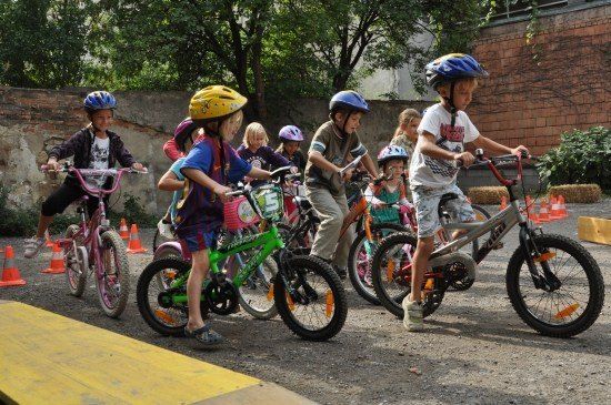 Eine Gruppe von Kindern mit Helmen fährt auf einem gepflasterten Bereich mit orangefarbenen Verkehrskegeln und einer Ziegelmauer im Hintergrund mit Fahrrädern.