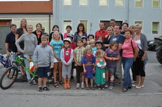 Eine Gruppe von Erwachsenen und Kindern, einige mit Helmen, stehen zusammen vor einem Gebäude mit Fahrrädern in der Nähe.
