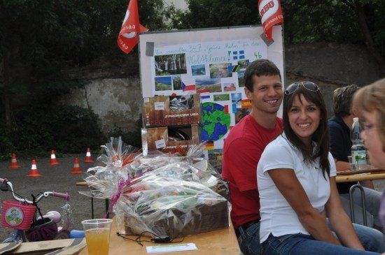 Ein Mann und eine Frau sitzen bei einer Outdoor-Veranstaltung zusammen, lächeln und sind von verschiedenen Gegenständen umgeben, einschließlich verpackter Geschenke und einem Fahrrad. Hinter ihnen ist ein Banner mit Bildern und Text angebracht, und im Hintergrund sind Verkehrskegel aufgestellt.