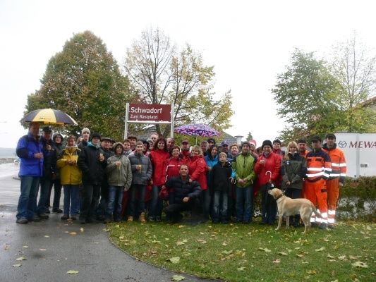 Eine Gruppe von Menschen, einige mit Regenschirmen, posiert für ein Foto vor einem Schild, das 'Schwadorf Zum Rastplatz' steht. Sie sind von Bäumen und Gras umgeben, mit einem Hund in der Nähe.