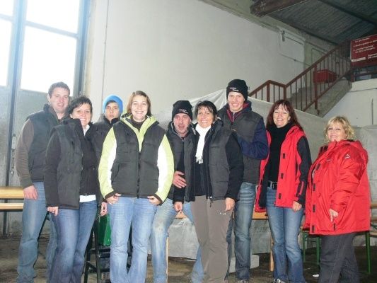 Eine Gruppe von Menschen in Winterjacken und Mützen posiert für ein Foto in einem Raum mit einer Treppe und einem Fenster.
