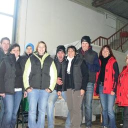 Eine Gruppe von Menschen in Winterjacken und Mützen posiert für ein Foto in einem Raum mit einer Treppe und einem Fenster.