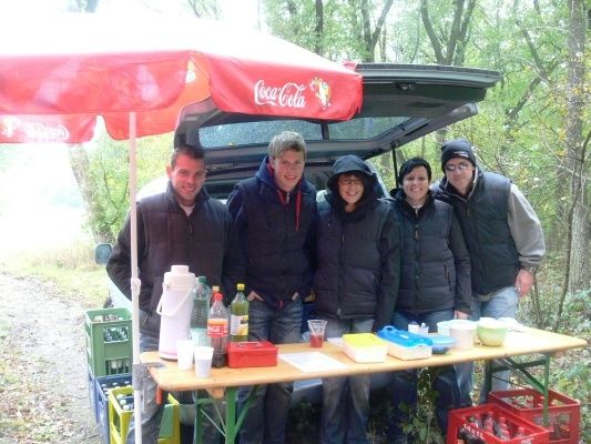 Fünf Personen stehen hinter einem Tisch mit Essen und Getränken, einer hält einen Coca-Cola-Regenschirm. Sie sind in Jacken gekleidet und stehen vor einem Auto mit offenem Kofferraum.