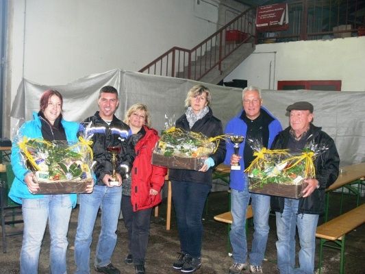 Eine Gruppe von Menschen, die Pokale und Blumensträuße halten, steht in einem Innenbereich. Hinter ihnen befindet sich eine Treppe und eine weiße Abdeckung.