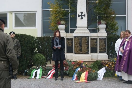 Eine Frau steht vor einem Denkmal, hält ein Stück Papier in der Hand, umgeben von Blumen, Flaggen und Militärangehörigen.