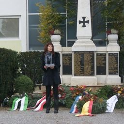 Eine Frau steht vor einem Denkmal, hält ein Stück Papier in der Hand, umgeben von Blumen, Flaggen und Militärangehörigen.