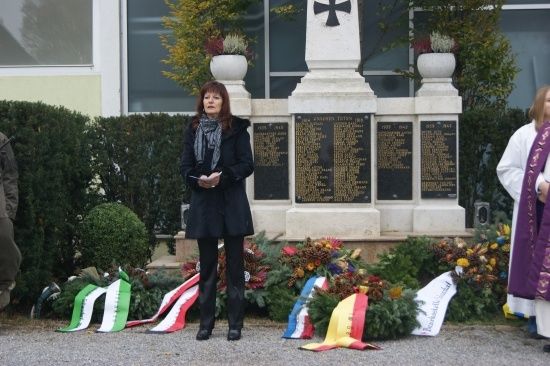 Eine Frau steht vor einem Denkmal mit einem Kreuz, umgeben von Kränzen und Flaggen, möglicherweise während einer Zeremonie.