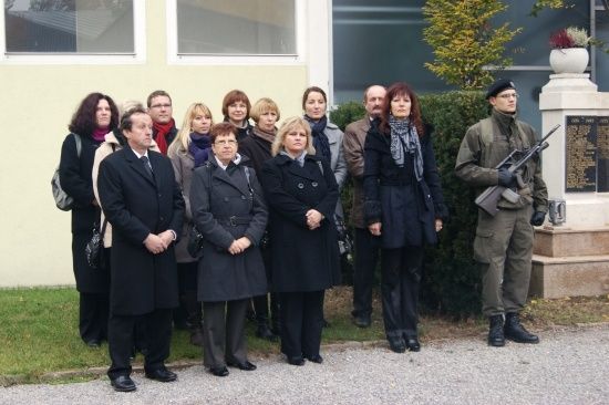 Eine Gruppe von Menschen in formeller Kleidung steht vor einem Gebäude mit einer Gedenktafel und einem Soldaten, der ein Gewehr hält.