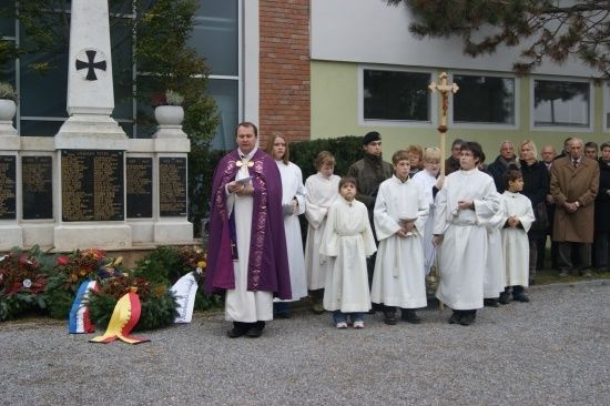 Ein Priester in einer violetten Robe steht vor einer Gruppe von Kindern in weißen Gewändern, alle versammelt vor einem Denkmal mit einem Kreuz und Blumen.